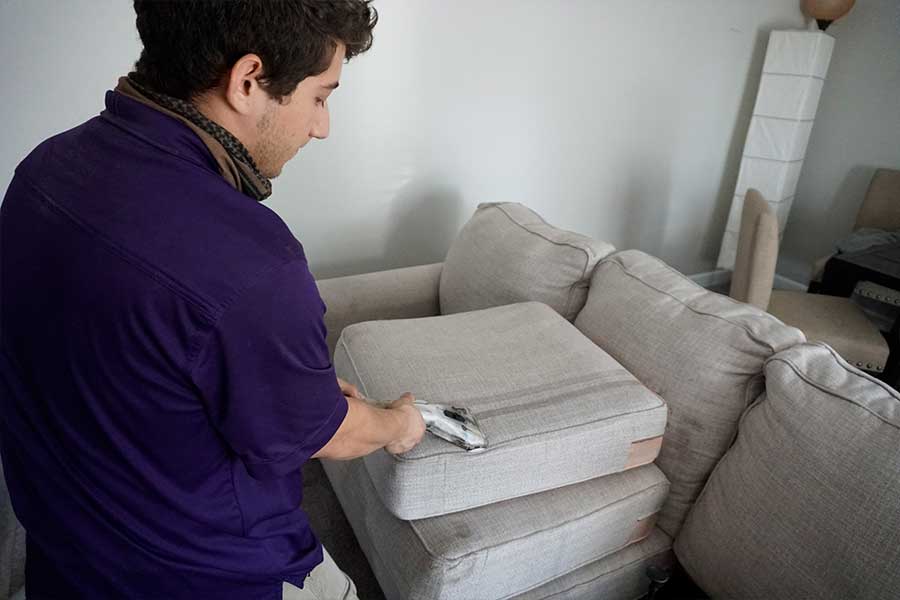 Man in purple shirt using upholstery cleaning service techniques to refresh a light gray fabric sofa, demonstrating spot cleaning with a cloth