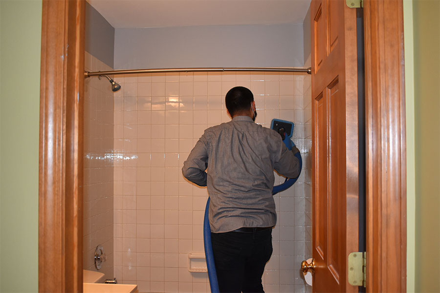 Worker from a bathroom tile and grout cleaning service meticulously scrubs white wall tiles in a shower with a blue handled brush, ensuring a sparkling clean finish