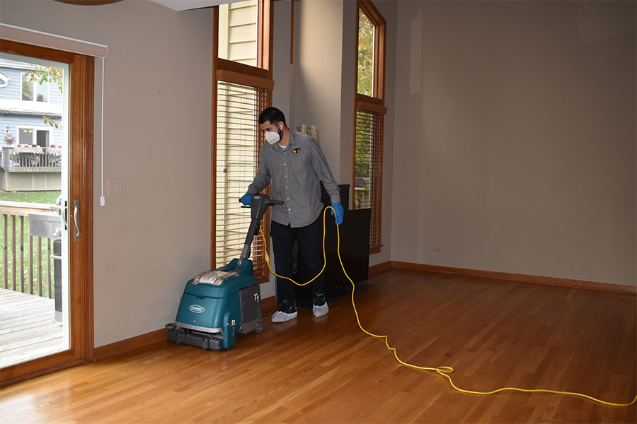 A dedicated worker from a wood floor cleaning service carefully maneuvers a floor machine over polished hardwood, in a bright room with tall windows and an open patio door