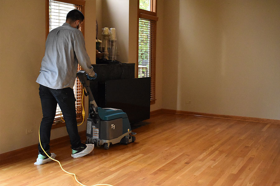 A technician from a professional hardwood floor cleaning service attentively operates a floor polishing machine across a gleaming wooden floor in a spacious room with natural light