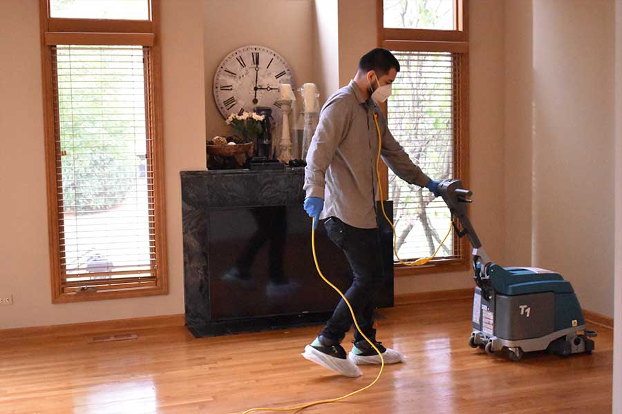 A professional from a hardwood floor cleaning service meticulously operates a floor buffing machine on a shiny wooden floor in a well-lit room with large windows