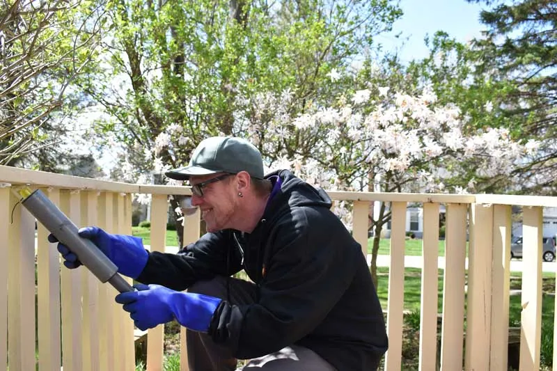 Smiling technician providing home pest control services, applying treatment along a wooden fence, with blooming trees in the background on a sunny day