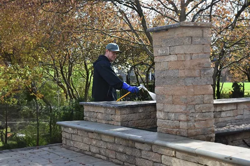 A pest control professional from a local service carefully applies treatment around the stone columns of an outdoor patio, ensuring a pest-free environment