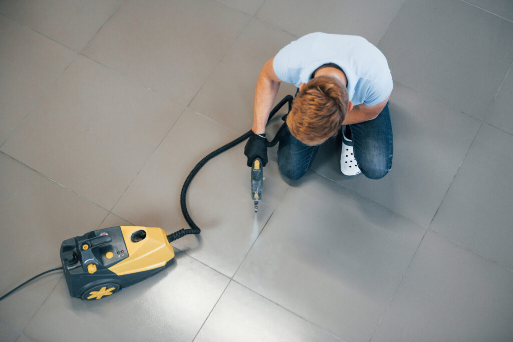 A man kneeling on a tiled floor using a steam cleaner to clean grout, demonstrating a common method used in tile and grout cleaning, helping homeowners understand how much grout cleaning costs.