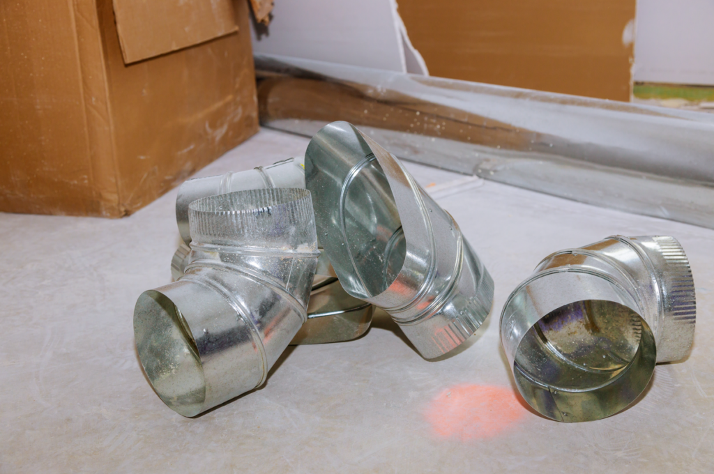 A collection of dirty metal duct components lying on the floor, including various angled connectors and sections, showing the importance of knowing the best ways to clean air ducts. 