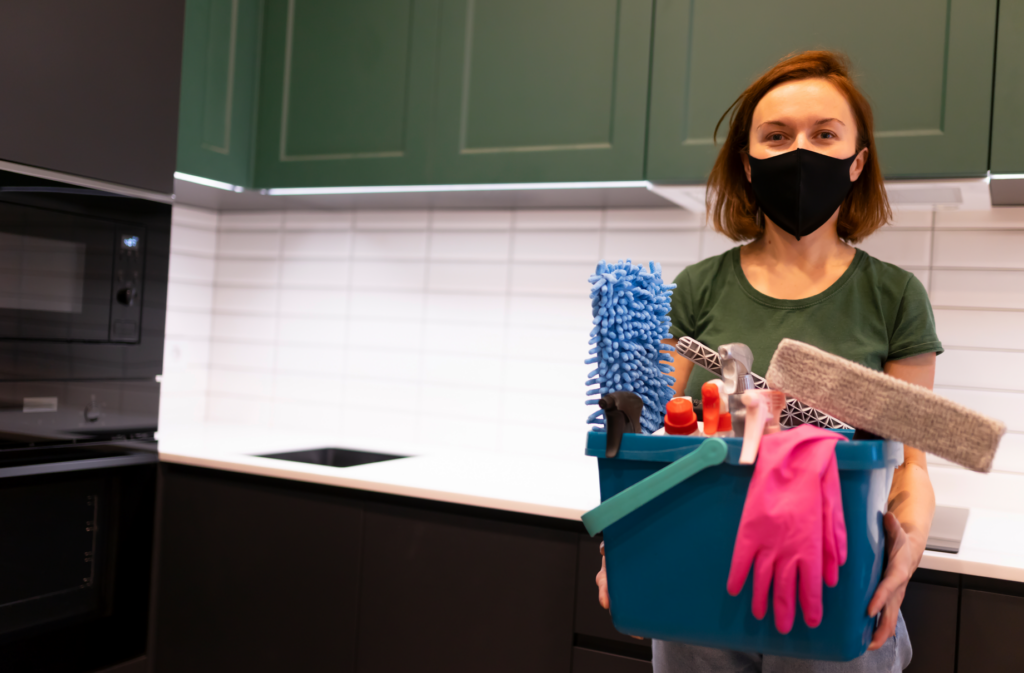 A person who knows the best ways to clean air ducts wearing a mask holding a bucket full of various cleaning supplies, including brushes, cleaning sprays, gloves, and cloths, highlighting the importance of being prepared with the right tools and protective gear for thorough air duct cleaning and maintaining a clean home environment.