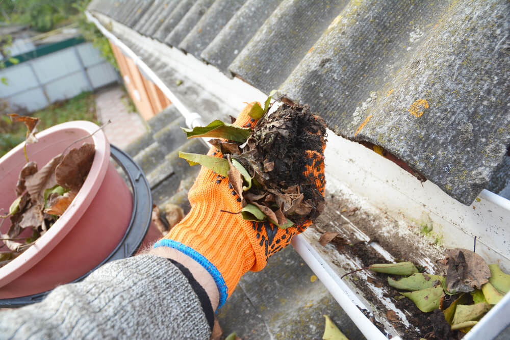 A person wearing orange gloves cleaning out leaves and dirt from a house gutter. This shows a typical task involved in home maintenance, which impacts how much gutter cleaning costs.