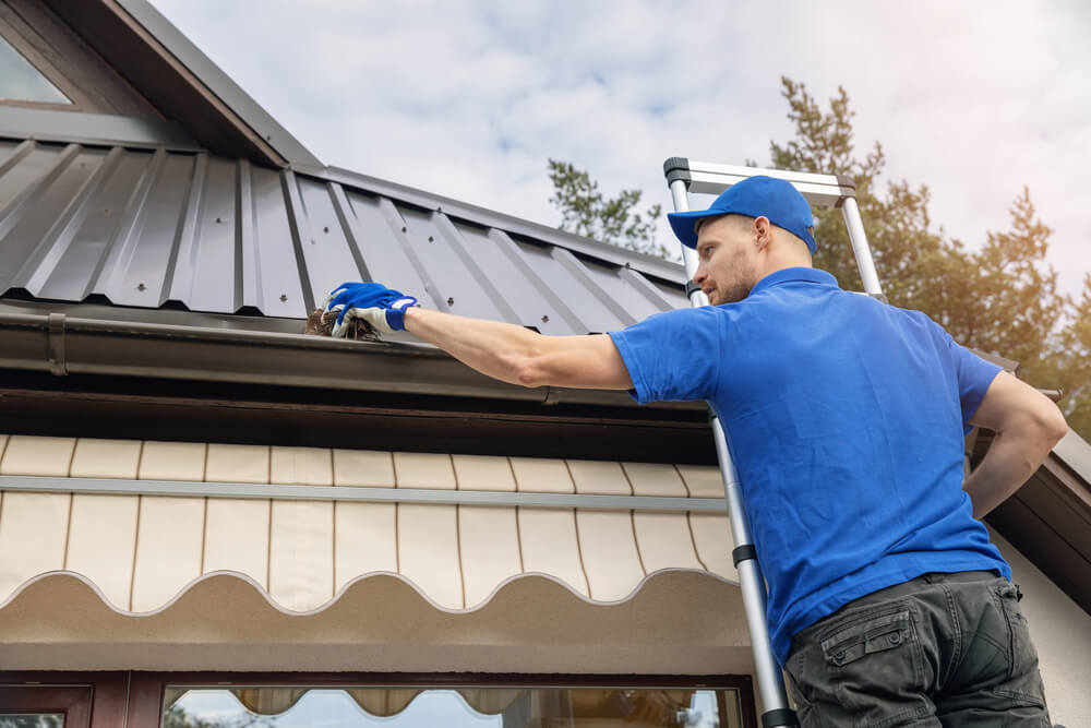 A professional in a blue uniform standing on a ladder cleaning debris from a house gutter. Hiring a professional for this service is a key factor when estimating  and answering the question - how much does gutter cleaning costs.