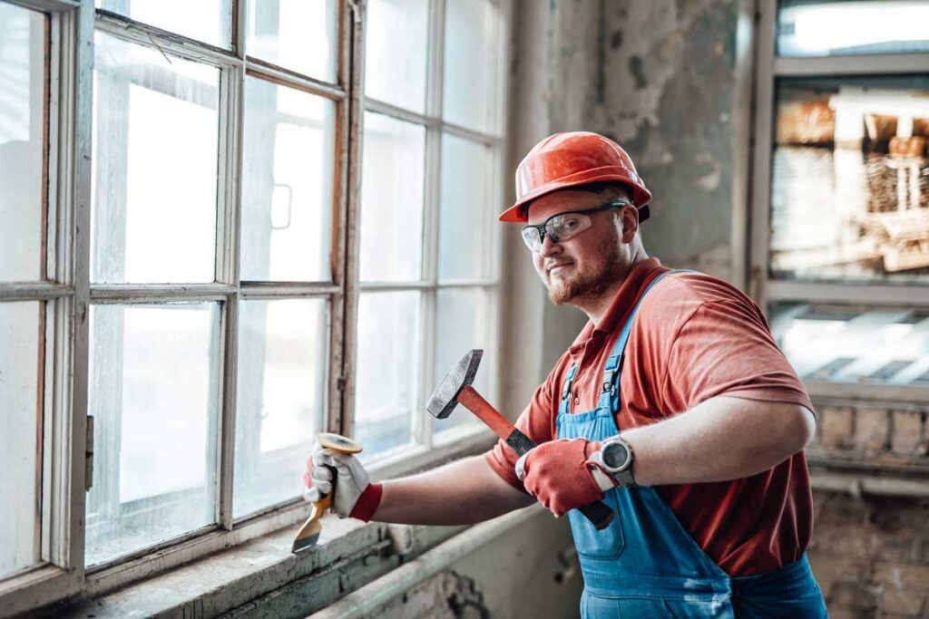 A construction worker wearing red safety gear and a hard hat carefully removes old wooden window frames with a hammer and scraper during a window replacement Chicago IL project inside a weathered, sunlit building.